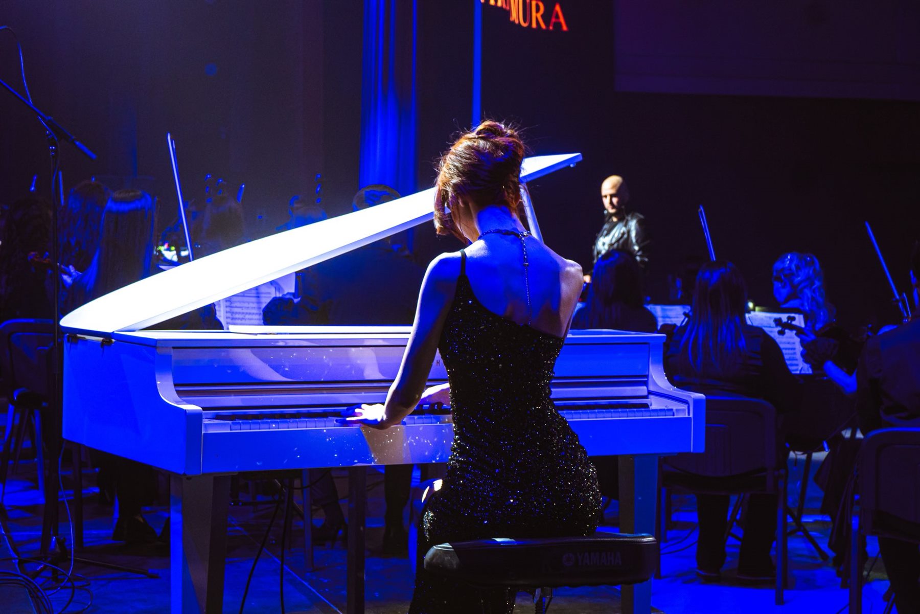 Arina Mura performing on grand piano on stage with full orchestra, blue stage lighting
